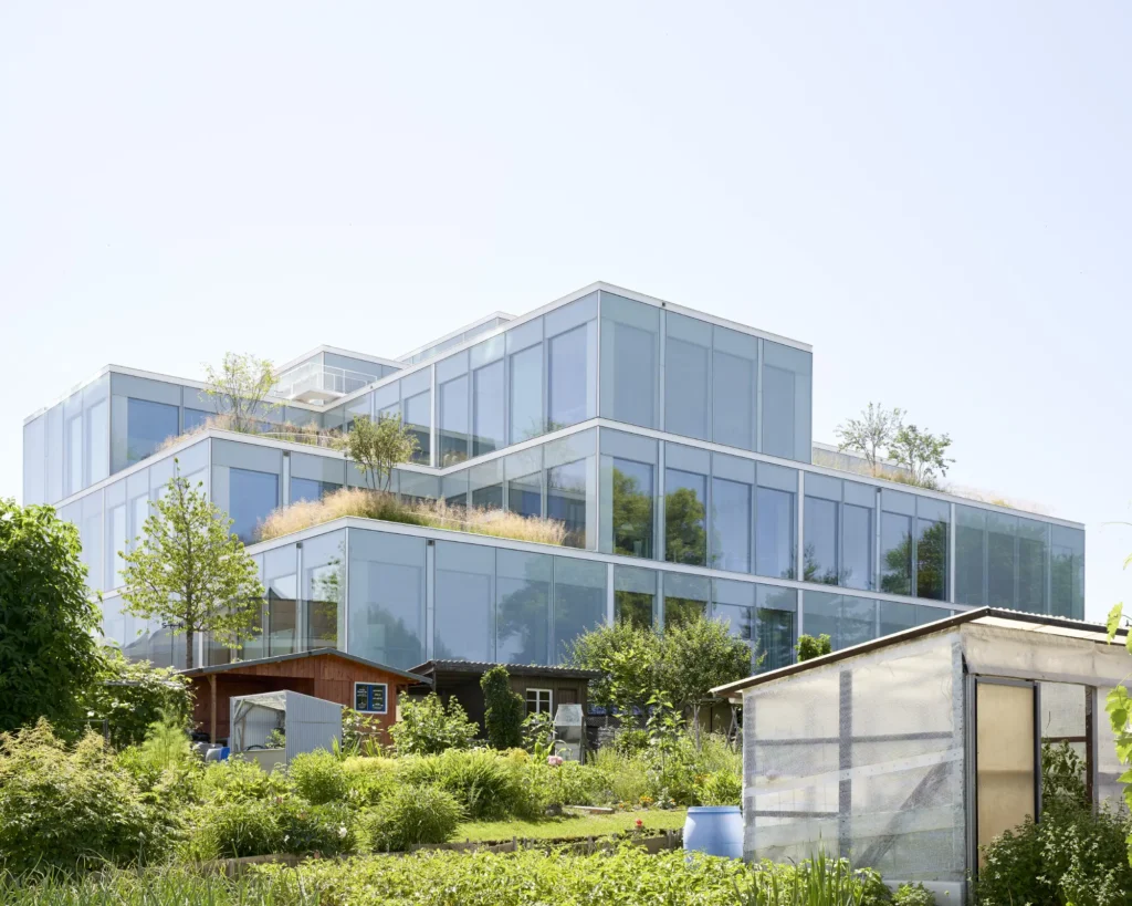 Stacked glass volumes of the SQUARE Learning Center seen through the urban gardens of the neighbourhood